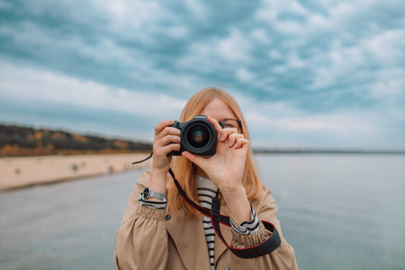 Photography and travel. Young woman in hat holding camera sitting on wooden fishing pier with...
