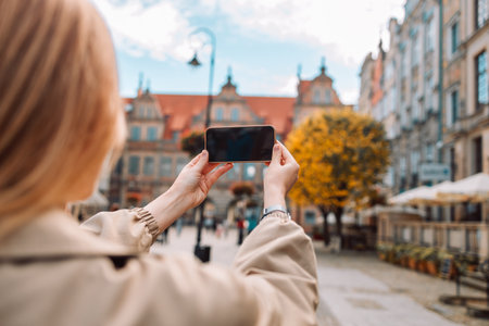 Happy young European lady tourist in hat makes photo or video on smartphone in old center of Gdansk, Poland, outdoor. Vacation tourism with app for travel blog, holiday journey, social media, active lifestyle, shooting vlog.の写真素材