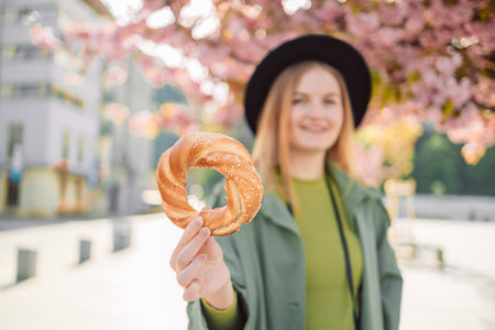 Happy young blonde female tourist with stylish clothes and a boho hat holds bagel obwarzanek traditional polish cuisine snack on blossoming sakura trees background in Krakow. Traveling Europe. High quality photoの写真素材