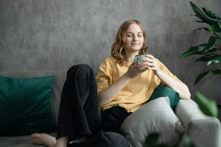 Close up of a beautiful happy girl sitting on couch drinking hot coffee and enjoying morning, being...