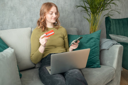 Young woman holding credit card and using laptop computer sitting on couch at home. Pretty relaxed lady holding smartphone buying in ecommerce shop, watching videos online, texting or checking social media news. High quality photoの写真素材