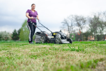 A young woman is mowing a lawn with a lawn mower in his beautiful green floral summer garden. A professional gardener with a lawnmower cares for the grass in the backyard. High quality photoの写真素材