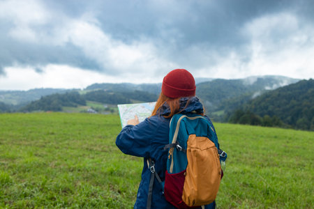 Stylish hipster 30s woman holding paper map, wearing backpack looking at mountain view while...