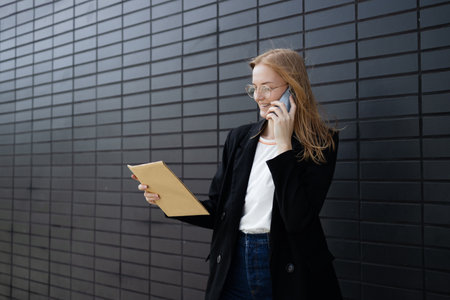 Portrait of corporate woman answers phone call while walking to the office building. Businesswoman calling someone, holding documents. High quality photoの写真素材