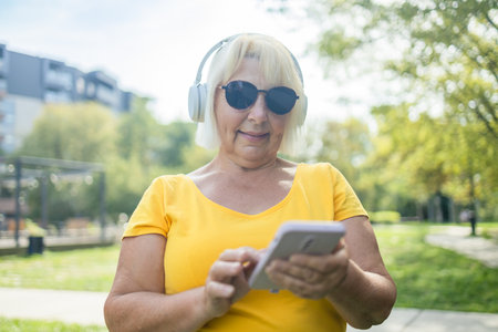Cheerful caucasian mature woman wife using smart phone cellphone, choosing sound track online while listening to the music in headphones in park. High quality photoの写真素材