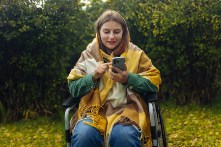 Young woman in bright stylish clothes with physical disability uses a mobile phone, reads news, communicates on social networks sitting in a wheelchair in park in autumn nature. High quality photoの写真素材