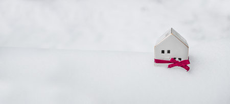 Merry Christmas and 2024 happy new year horizontal banner with small toy model house wrapped in red satin ribbon on winter snow background. Miniature white toy house model. High quality photoの写真素材