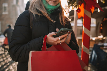 Cheerful smiling woman typing on telephone at Christmas market background. New Years is soon. Winter holidays, preparation and shopping conceptの写真素材