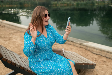 Photo of cute funny brunette lady making a selfie or video with front camera on mobile phone, girl having a funny video call and laughing in the park during break in university, wifi in public placesの写真素材