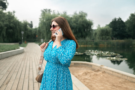 Young pretty woman model in stylish dresses talk by mobile cell phone look aside standing on summer in spring park outdoors. Urban lifestyle concept. High quality photoの写真素材