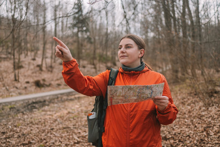 Young tourist girl in sports clothes with a map in her hands shows the directions of the path and looking around standing in the autumn forest. Hiking and navigation concept. High quality photoの写真素材