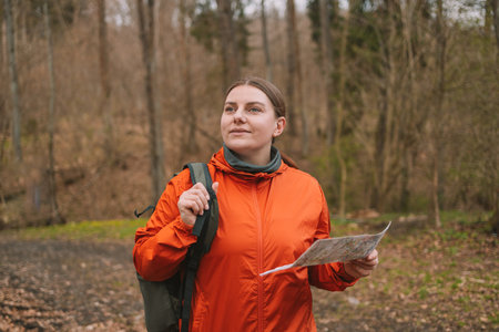 Young tourist girl in sports clothes with a map in her hands shows the directions of the path and looking around standing in the autumn forest. Hiking and navigation concept. High quality photoの写真素材