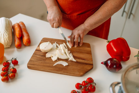 Senior housewife slices onion into pieces on a wooden chopping board in a modern kitchen. Female cutting fresh raw onion on wooden background. High quality photoの写真素材