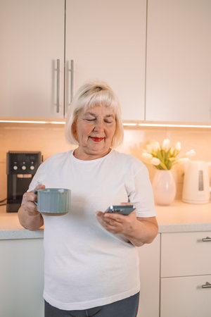 Middle aged woman drinking fresh morning coffee and text messaging while relaxing at home in her kitchenの写真素材