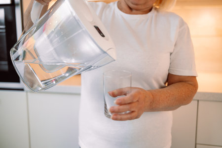 Smiling senior woman pouring filtered water into glass, kitchen interior. Attractive older woman in casual outfit holding glass of fresh water, copy space. High quality photoの写真素材