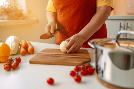 Female chef is precisely slicing fresh onions on wooden board while preparing italian pasta with vegetables and soup in kitchenの写真素材