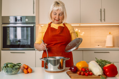 Beautiful happy senior woman is cooking in the home kitchen and testing some soup with fresh vegetables from the pan on the stove. High quality photoの写真素材