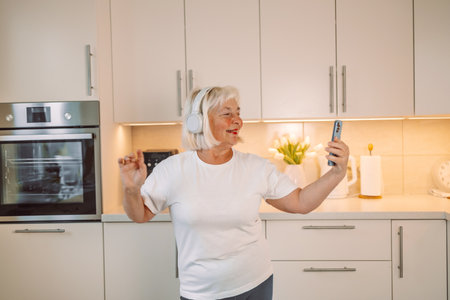 60s happy woman is using mobile phone standing in the kitchen. High quality photoの写真素材