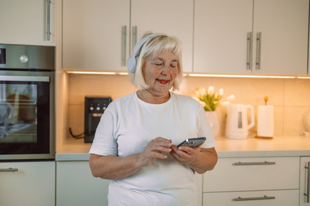 60s happy woman is using mobile phone standing in the kitchen. High quality photoの写真素材