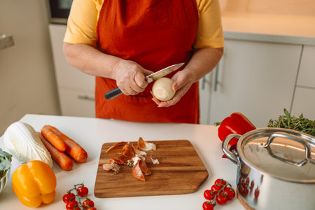 Female chef is precisely slicing fresh onions on wooden board while preparing italian pasta with vegetables and soup in kitchenの写真素材