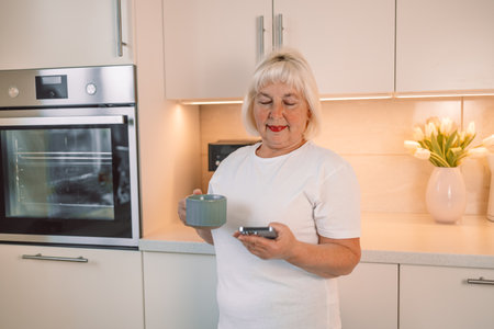 Middle aged woman drinking fresh morning coffee and text messaging while relaxing at home in her kitchenの写真素材