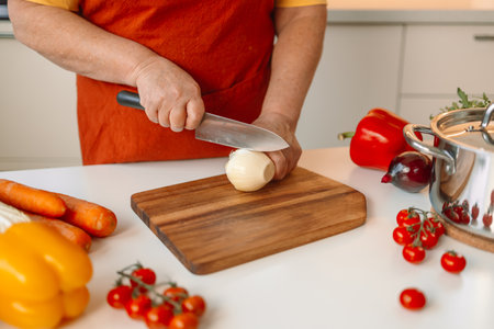 Female chef is precisely slicing fresh onions on wooden board while preparing italian pasta with vegetables and soup in kitchenの写真素材