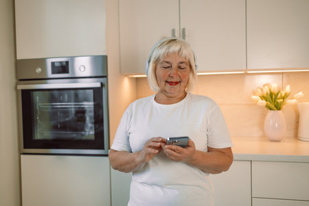 60s happy woman is using mobile phone standing in the kitchen. High quality photoの写真素材