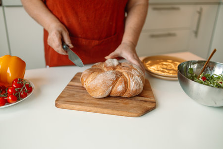 Caucasian woman cutting bread on wooden board. Bakehouse. Bread production. Cooking breakfast. Hand with a knife, woman cuts bread with a knife. High quality photoの写真素材