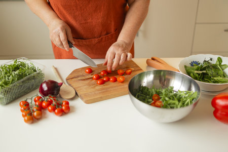 Female chef is precisely slicing fresh cherry tomato on wooden board while preparing Italian salad with vegetables in kitchenの写真素材