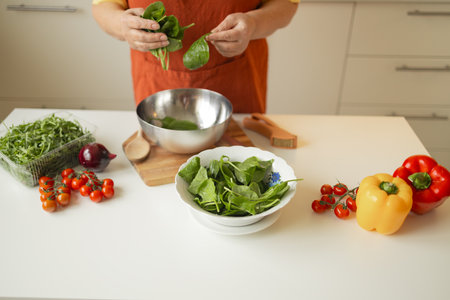 Senior 60s blond Caucasian smiling woman holding a knife and fresh spinach whole preparing a bowl of healthy diet salad in her rustic eco kitchen. High quality photoの写真素材