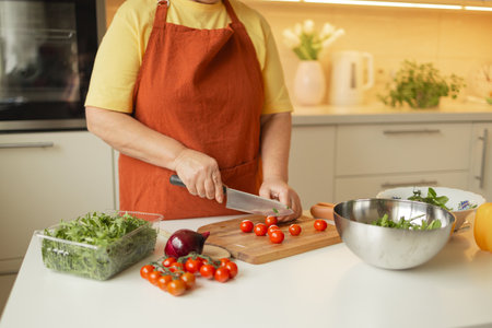 Female chef is precisely slicing fresh cherry tomato on wooden board while preparing Italian salad with vegetables in kitchenの写真素材