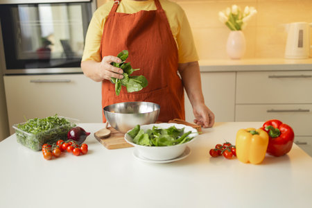 Senior 60s blond Caucasian smiling woman holding a knife and fresh spinach whole preparing a bowl of healthy diet salad in her rustic eco kitchen. High quality photoの写真素材