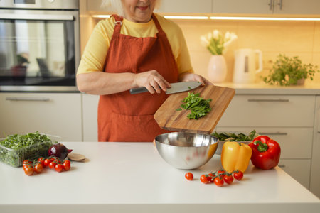 Senior 60s blond Caucasian smiling woman holding a knife and fresh spinach whole preparing a bowl of healthy diet salad in her rustic eco kitchen. High quality photoの写真素材