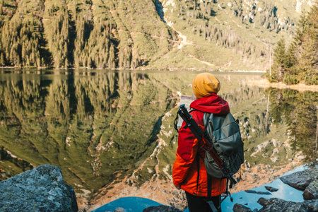 Shot of back view of senior 50s woman in red jacket standing on top of mountain and looking at Morskie oko lake. Female with backpack on mountain edge and enjoying the beautiful view in Poland, Tatryの写真素材