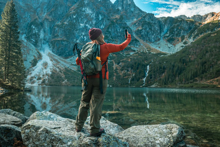Rear shot of traveler woman influencer in sportswear with backpack standing on the rocky shore and making selfie on smart phone. Taking photo of green hills and mountains on Morskie Oko lake. Hiking travel and adventure in Poland, Tatryの写真素材