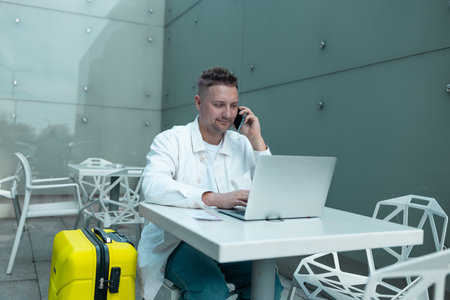 Happy traveling Caucasian man in stylish clothes uses mobile smartphone and laptop, checking trip destination on Internet. Male waiting at flight gates for plane boarding, smiling have a vacation trip. High qualityの写真素材