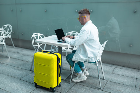 Happy traveling Caucasian man in stylish clothes uses mobile smartphone and laptop, checking trip destination on Internet. Male waiting at flight gates for plane boarding, smiling have a vacation trip. High qualityの写真素材
