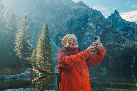 Rear shot of traveler 60s woman influencer in sportswear with backpack standing on the rocky shore and making selfie on smart phone. Taking photo of green hills and mountains on Morskie Oko lake. Hiking travel and adventure in Poland, Tatryの写真素材