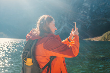 Rear shot of traveler woman influencer in sportswear with backpack standing on the rocky shore and making selfie on smart phone. Taking photo of green hills and mountains on Morskie Oko lake. Hiking travel and adventure in Poland, Tatryの写真素材