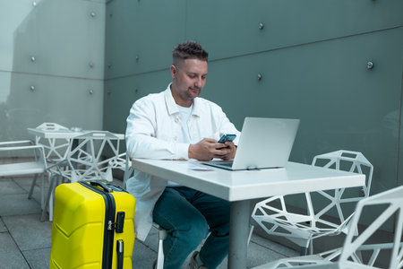 Young Caucasian male passenger in stylish casual clothes using smartphone and laptop keyboard, sit has training online meeting on laptop computer in airline lounge wait transit international airport.の写真素材