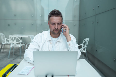 Happy traveling Caucasian man in stylish clothes uses mobile smartphone and laptop, checking trip...