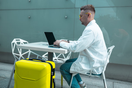 Happy traveling Caucasian man in stylish clothes uses mobile smartphone and laptop, checking trip destination on Internet. Male waiting at flight gates for plane boarding, smiling have a vacation trip. High qualityの写真素材