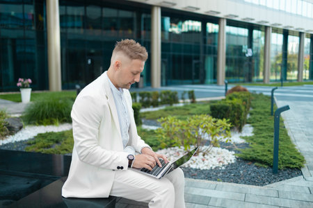 Focused young businessman corporate worker investor with suitcase sitting near business centre waiting, working remote on laptop. High qualityの写真素材