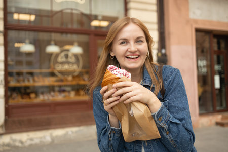 Smiling European girl woman tourist enjoying breakfast croissant near city cafe. Caucasian lady female smelling relaxing outdoors. High qualityの写真素材
