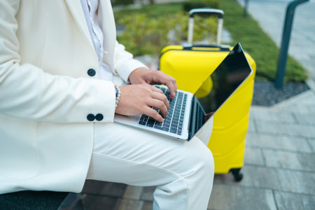 Focused young businessman corporate worker investor with suitcase sitting near business centre waiting, working remote on laptop. High qualityの写真素材