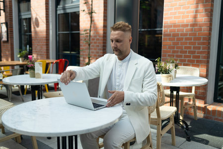 Happy successful European businessman using a laptop, talking and smiling outdoors sits at city cafe on city street outside. High qualityの写真素材