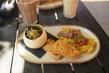 Platter with fried egg with greens, avocado, fried bacon, white bread croutons and pasta. A plate with a classic continental breakfast, standing on a wooden table in the restaurant by the window.の写真素材