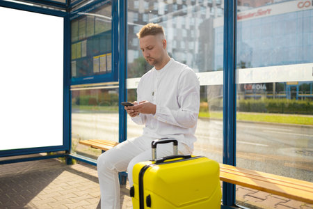Tourist Caucasian business man traveler with bag and suitcase sit at public transport bus station waiting with mobile smart phoneの写真素材