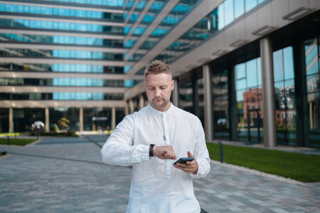 Focused young corporate worker investor businessman walking and using mobile phone waits for a client and looks at the watch on his hand outdoors near a business centre. High quality FullHD footageの写真素材
