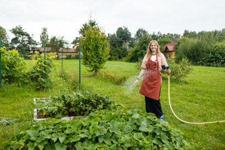 30s woman watering fresh plants growing at home vegetable garden. Gardener taking care of plants at the backyard of her house. Concept of sustainability and growing organic. High quality photoの写真素材